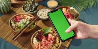 夏威夷戳碗，拿着彩色绿色屏幕的智能手机特写。由切成片的蔬菜、海鲜和绿叶植物制成的熟戳。健康的素食。亚洲素食生食，筷子