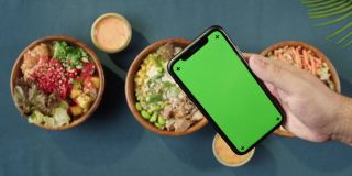 夏威夷戳碗，手持智能手机与彩色绿色屏幕顶部视图。由切成片的蔬菜、海鲜和绿叶植物制成的熟戳。健康的素食。亚洲素食生食，筷子