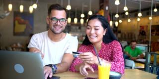 多种族的年轻夫妇一起在线购物的笔记本电脑，从现代和舒适的咖啡店