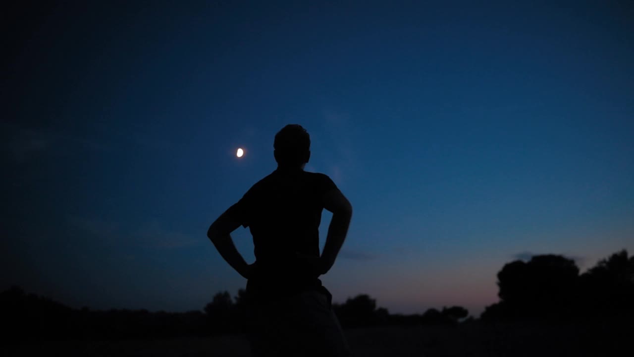 一个男人的剪影和乡村星空下年轻的月亮。