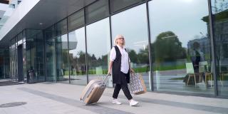 描绘活跃的老年商务女性游客带着行李箱走在城市背景的现代建筑上的老年女性