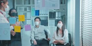 一群亚洲商人和妇女戴着面具，在办公室开会。员工集思广益，团队合作，计划和讨论项目。现代同事或同事的联合。