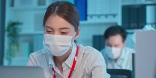 亚洲年轻的女商人戴着面具，在办公室里工作很累。男女职工在工作场所使用电脑加班至深夜，保持距离以防新冠病毒。