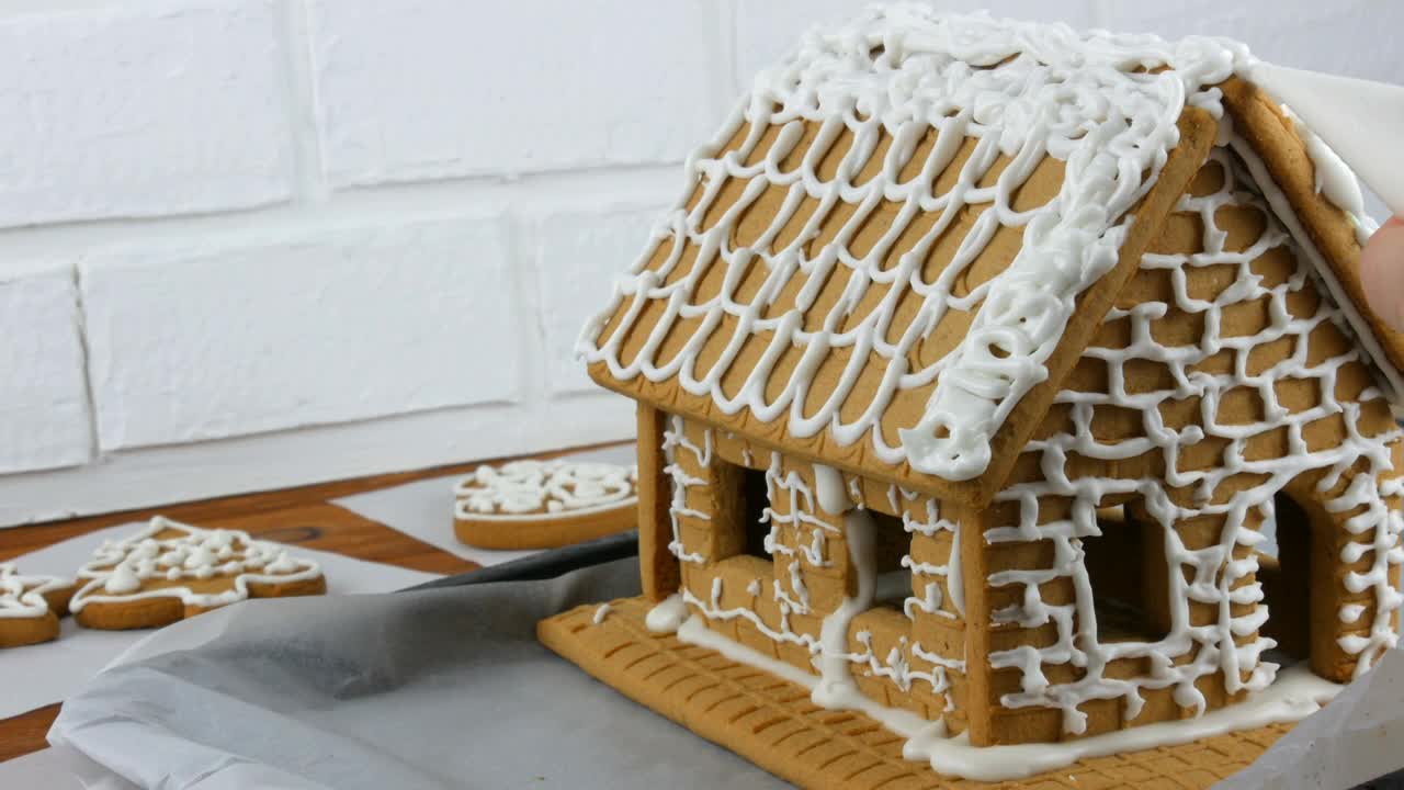 女人装饰姜饼饼干屋顶房子与糕点袋白糖糖霜，手在白色砖的背景。烹饪，自制姜饼屋圣诞假期。新年传统