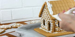 女人装饰姜饼饼干屋顶房子与糕点袋白糖糖霜，手在白色砖的背景。烹饪，自制姜饼屋圣诞假期。新年传统