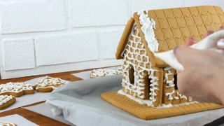 女人装饰姜饼饼干屋顶房子与糕点袋白糖糖霜，手在白色砖的背景。烹饪，自制姜饼屋圣诞假期。新年传统视频素材模板下载