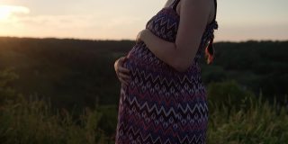 日落前，一个快乐的孕妇在高高的草丛中抚摸着自己的肚子。慈爱的母亲用手抚摸着大肚子。怀孕，母性，人，期望，夏天的概念