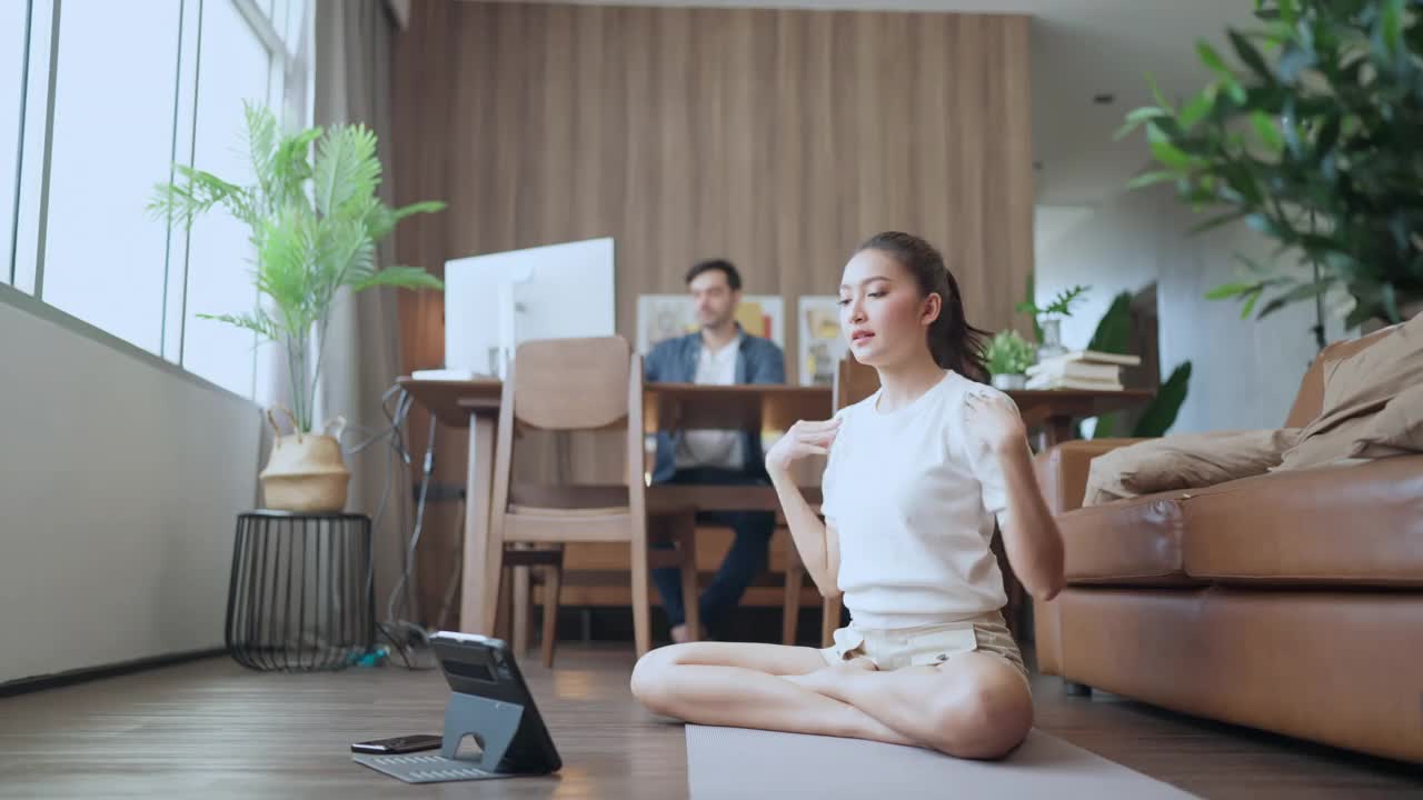 年轻的亚洲女性妻子训练瑜伽锻炼锻炼练习feomr在线课程在客厅与丈夫工作在后面，工作在家里呆在家里的状态秩序概念