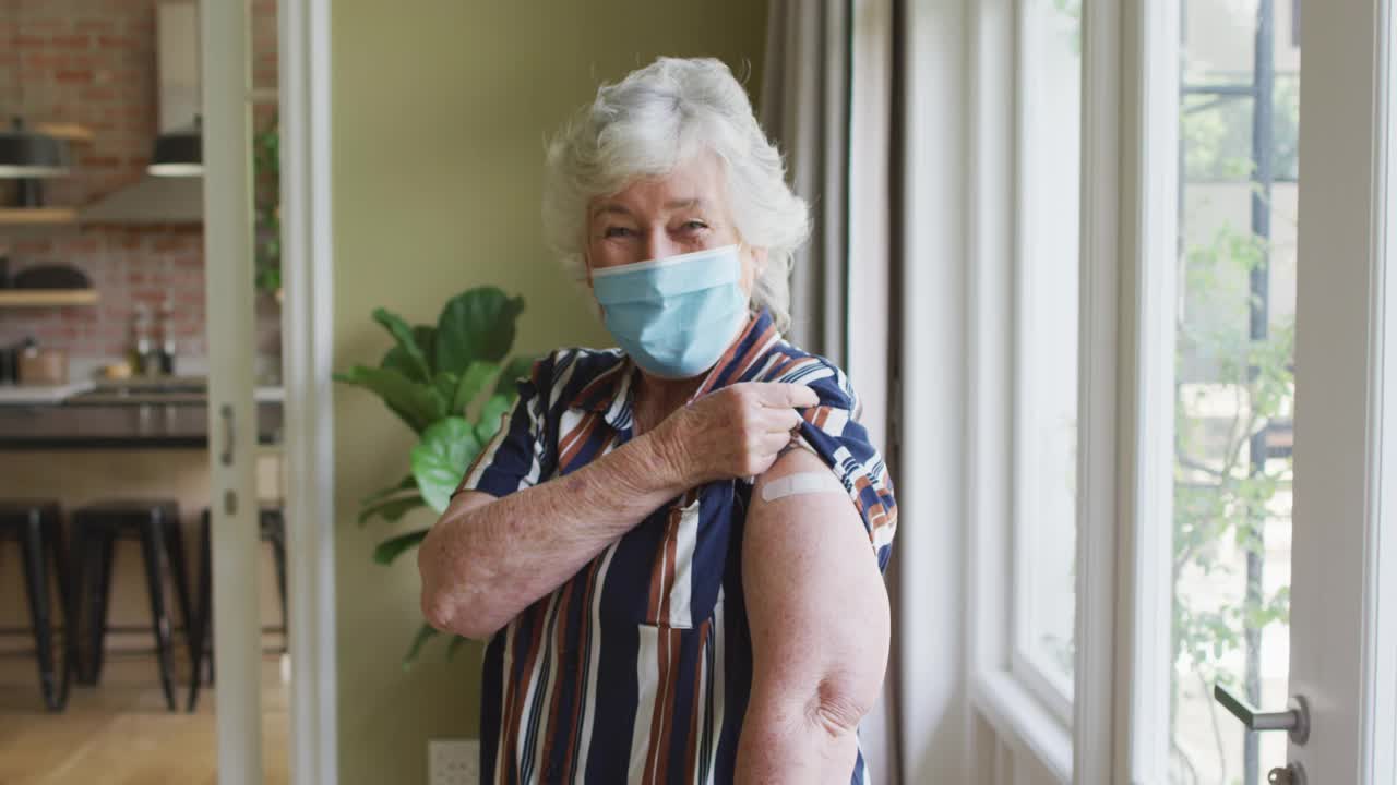 一名白人老年妇女戴着口罩，向人们展示她接种过疫苗的肩膀