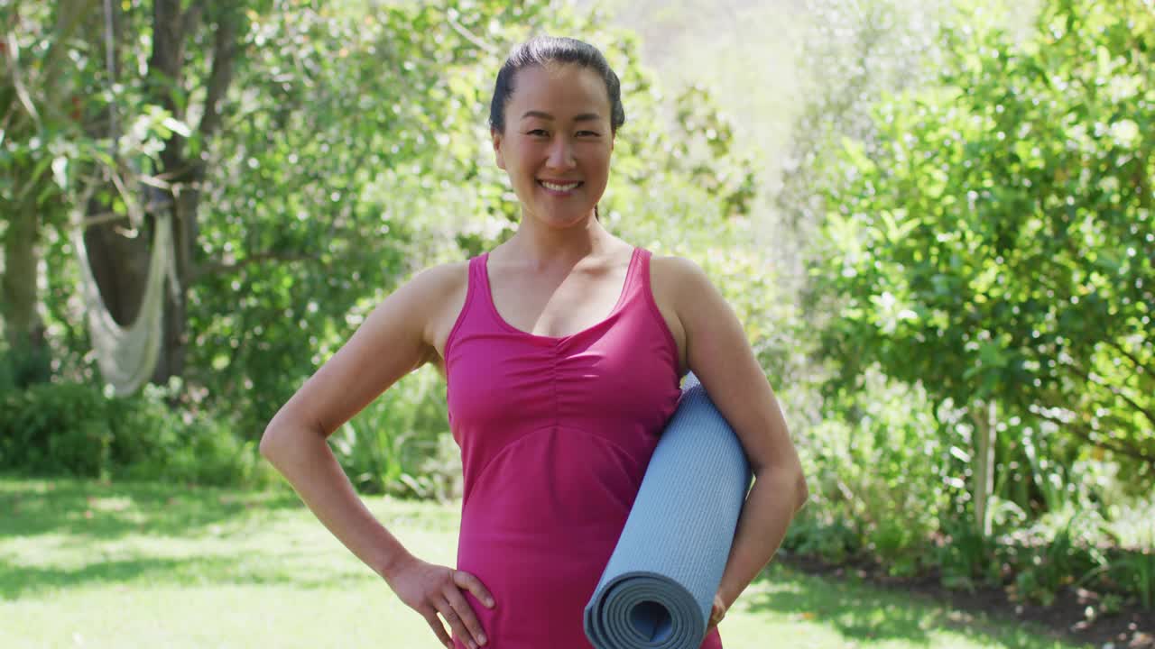 亚洲妇女的肖像，举着瑜伽垫微笑，站在公园
