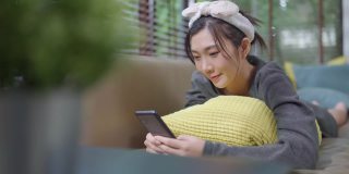 幸福快乐微笑年轻的成年亚洲女性女人穿着黄色的布手在智能手机交谈在家里的沙发远程隔离锁定时期家内部背景