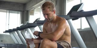 英俊的肌肉男坐在跑步机上跑步，通过智能手表查看时间，在健身房里喝水，完成了锻炼。赤裸上身的运动男在跑步机上锻炼。有氧运动训练