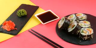阴阳寿司卷上有鳗鱼和红色的鱼鲑鱼，时髦的珊瑚黄色背景板，黑色的板，绿色的芥末和粉红色的姜。极简主义风格的日本料理