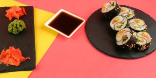 黑色的木条以阴阳鱼寿司卷为原料，搭配鳗鱼和红色的鲑鱼，时髦的珊瑚黄色背景板，黑色的板，绿色的芥末和粉红色的姜。极简主义风格的日本料理