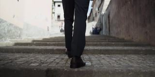背面的男人穿着优雅的西装和黑色的鞋子沿着古老的街道爬混凝土楼梯，特写的人的脚爬慢动作楼梯。在街上与人擦肩而过的概念