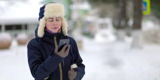 一位戴着帽子、戴着耳罩、穿着夹克的年轻女子的肖像，她在电话里不停地打电话。背景是白雪覆盖的街道