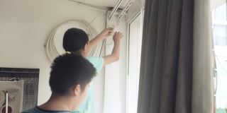 亚洲女孩帮妈妈在家里的窗户旁挂新床单白窗帘，隔离生活理念。