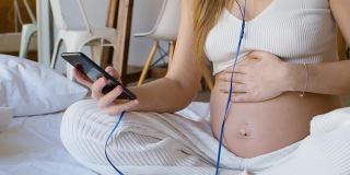一个年轻的孕妇坐在床上，耳机里戴着智能手机。她积极地在网上交流——通过她的智能手机写信和发送信息