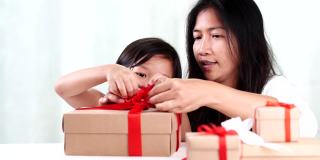 年轻的亚洲母亲和女儿帮助包装乡村礼品盒礼物领带红色丝带蝴蝶结。赠送的季节，圣诞节，新年快乐，生日，情人节，周年纪念日，母亲节。