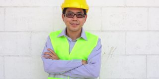 亚洲男性工人微笑着看着相机，双臂交叉，自信的劳动快乐在建筑工地，职业就业