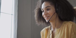 微笑的非裔美国妇女戴着耳机使用笔记本电脑，交谈，工作客户支持服务操作员在家里的办公室，女孩戴着耳机麦克风