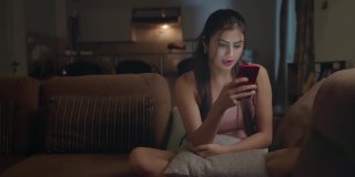 印度亚洲美丽的现代女性坐在沙发上，微笑着在智能手机上阅读短信