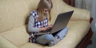 女孩坐在家里的沙发上，热情洋溢地在笔记本电脑上打字。她做家庭作业。