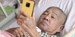 一位亚裔华裔老人躺在医院病房的病床上，用智能手机与家人视频通话