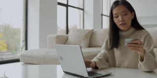 一个年轻的亚洲女人拿着信用卡，用着笔记本电脑。一位在家工作的女企业家。网上购物，在家工作的概念