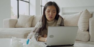 一个亚洲女人用餐巾擤鼻涕，坐在家里的办公桌旁，用笔记本电脑学习，一个生病的女人得了流感，用毯子盖住自己，得了感冒