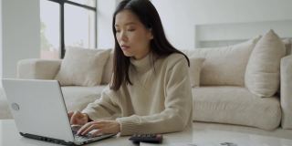 亚洲女人带着笔记本电脑和计算器在客厅里。一位认真的女士在笔记本电脑的帮助下计算房子的财务状况