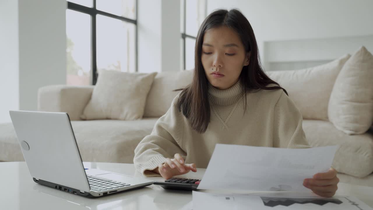 一个亚洲女人在客厅里拿着笔记本电脑和计算器。一位认真的女士在笔记本电脑的帮助下计算家里的财务状况，并查看她的账单