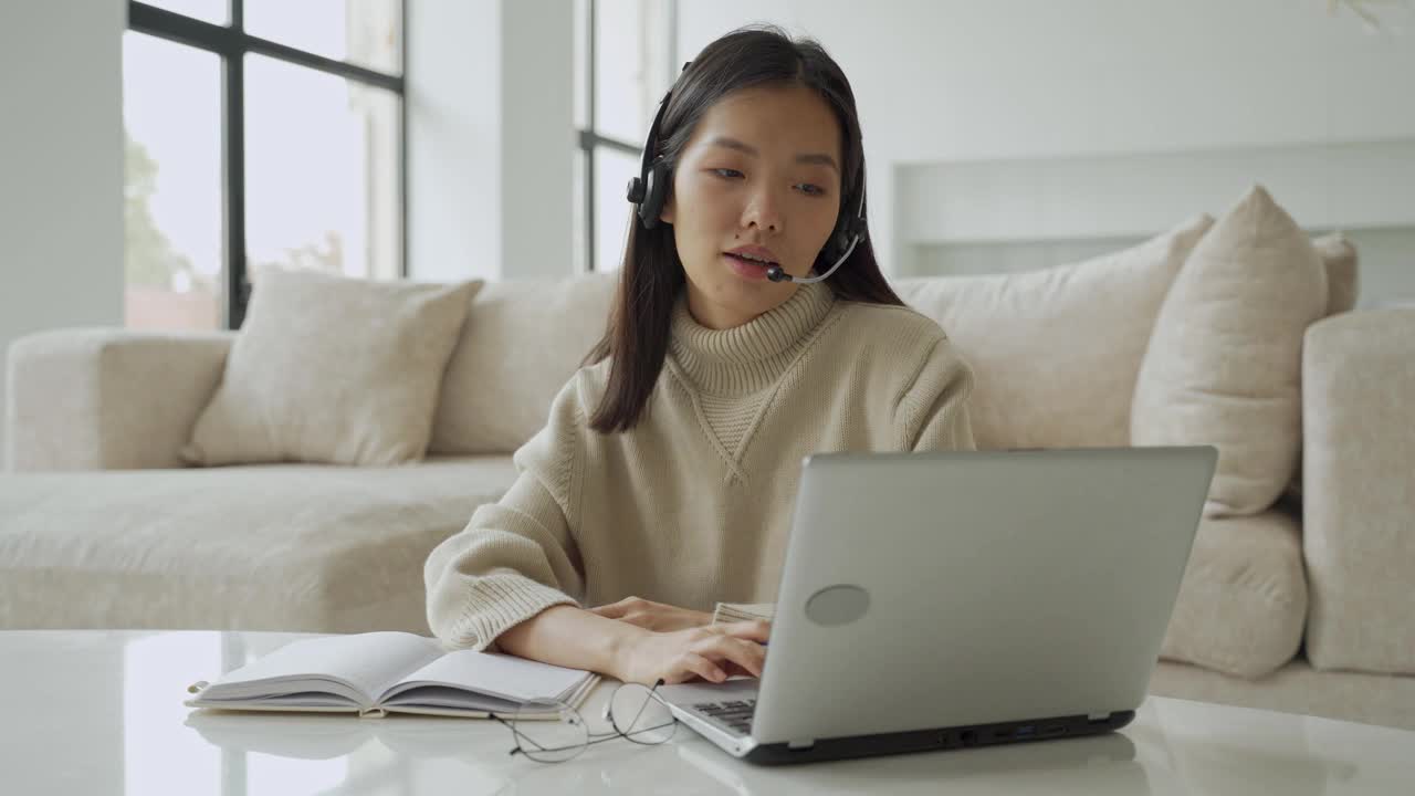 一位亚洲女性戴着耳机看着笔记本电脑屏幕，与对话者交流。韩国女商人戴着耳机视频呼叫客服