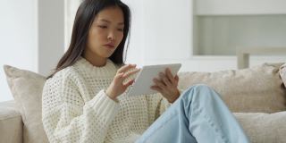 一位亚洲妇女坐在客厅的沙发上使用平板电脑。一个快乐的女人在家里用平板电脑看视频和上网