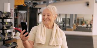 在健身房描绘老妇人。老年人的体育文化。培训和个人课程。快乐的人微笑着，在手机上拍照，视频通话随着音乐起舞