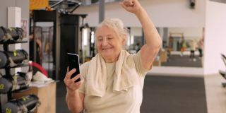 在健身房描绘老妇人。老年人的体育文化。培训和个人课程。快乐的人微笑着，在电话里拍照，视频通话。运动就是力量