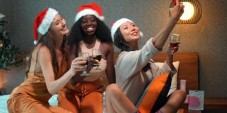 年轻的女孩来自欧洲、亚洲和非洲，穿着红色的新年衣服，戴着红色的帽子，坐在圣诞树前的床上，手里拿着酒杯，用手机拍照，微笑着庆祝圣诞节