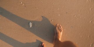 年轻人在沙滩上踩着沙子的观点。一个人在热带沙滩上度假的第一人称视角。男性穿过海洋海滩的波浪。