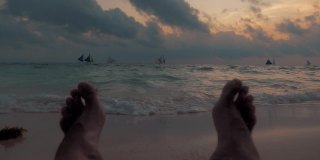 两名男性的脚在日落的沙滩上放松的POV视图。一个男人躺在沙滩上。看日落。美丽的日落。旅行的概念。慢动作镜头。长滩岛,菲律宾。