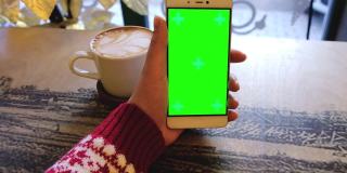 身穿红色圣诞毛衣的女性手持绿色屏幕手机和一杯咖啡放在白色的桌子上。女人使用带有色度键的手机。上网，看内容，轻按中央屏幕
