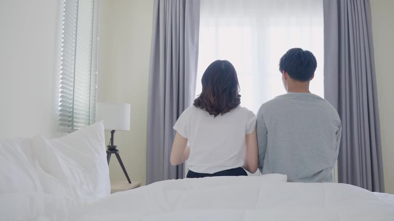 亚洲夫妇早上醒来后伸出手的背影。迷人的新婚男女在清晨一起从卧室的床上起床后，穿着睡衣感到快乐和放松。