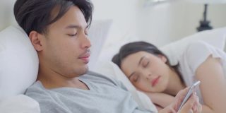 睡在床上的亚洲妻子试图偷看丈夫的手机。迷人有趣的夫妇躺在卧室里，年轻的男性打字短信，而女性女朋友在家里睡觉。