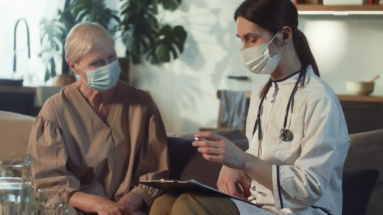 家访时，身穿白大褂的年轻女医生向戴口罩的老年妇女展示装有疫苗的药瓶。