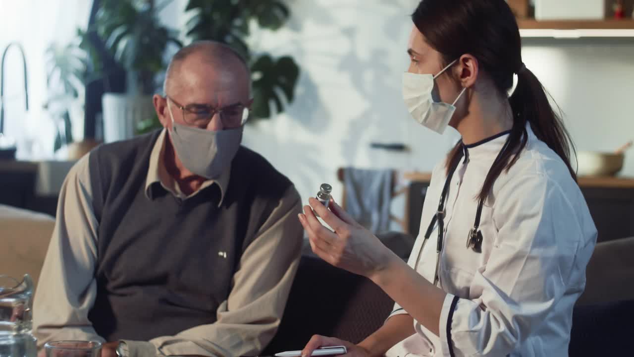 年轻漂亮的女医生向65-70岁戴着医用口罩的快乐老人展示装有疫苗的烧瓶