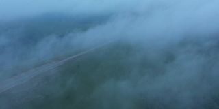 从山顶上可以看到穿过山丘的山路。无人机在绿色田野上空的云层中飞行