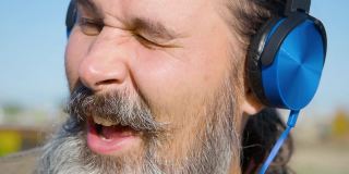 留着胡子的中年男子戴着耳机听着音乐，跟着音乐唱歌，并随着音乐的节拍点头。面部特写