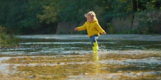 一个可爱的小女孩穿着黄色的胶靴在河上走着。缓慢的运动。跟随拍摄