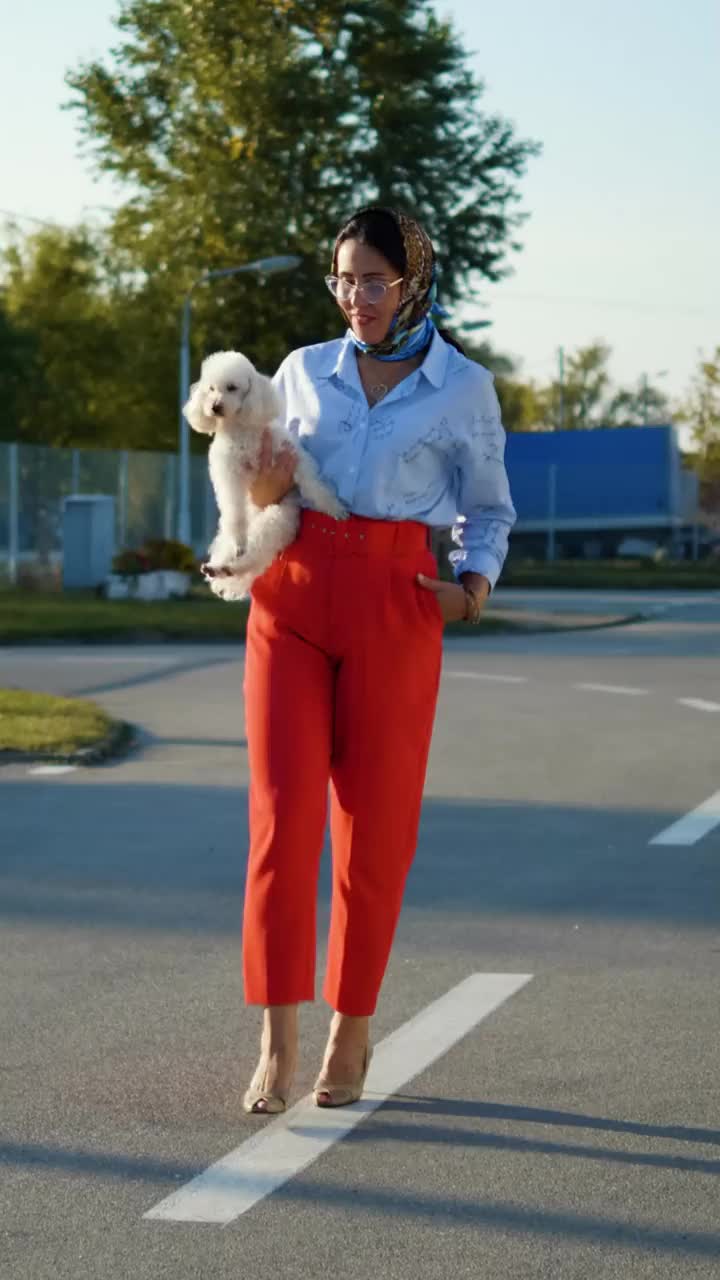 一个漂亮的中年女人抱着一只小马耳他犬走在街上。现代女性带着宠物在城市里散步。垂直