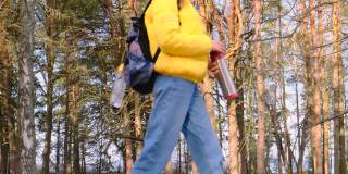 搭便车旅行的概念。一名年轻的女游客背着黄色夹克的背包，手里拿着保温瓶，沿着松树林走着。慢镜头,4 k
