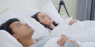 亚洲新婚夫妇躺在床上，丈夫使用手机。漂亮的年轻男人和女人在睡衣享受清晨的活动在卧室在家里。家庭关系的概念。